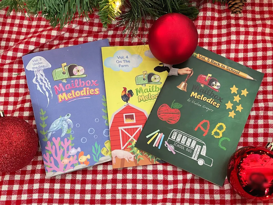 Three Mailbox Melodies music booklets arranged with red ornaments and pine garland as a holiday gift