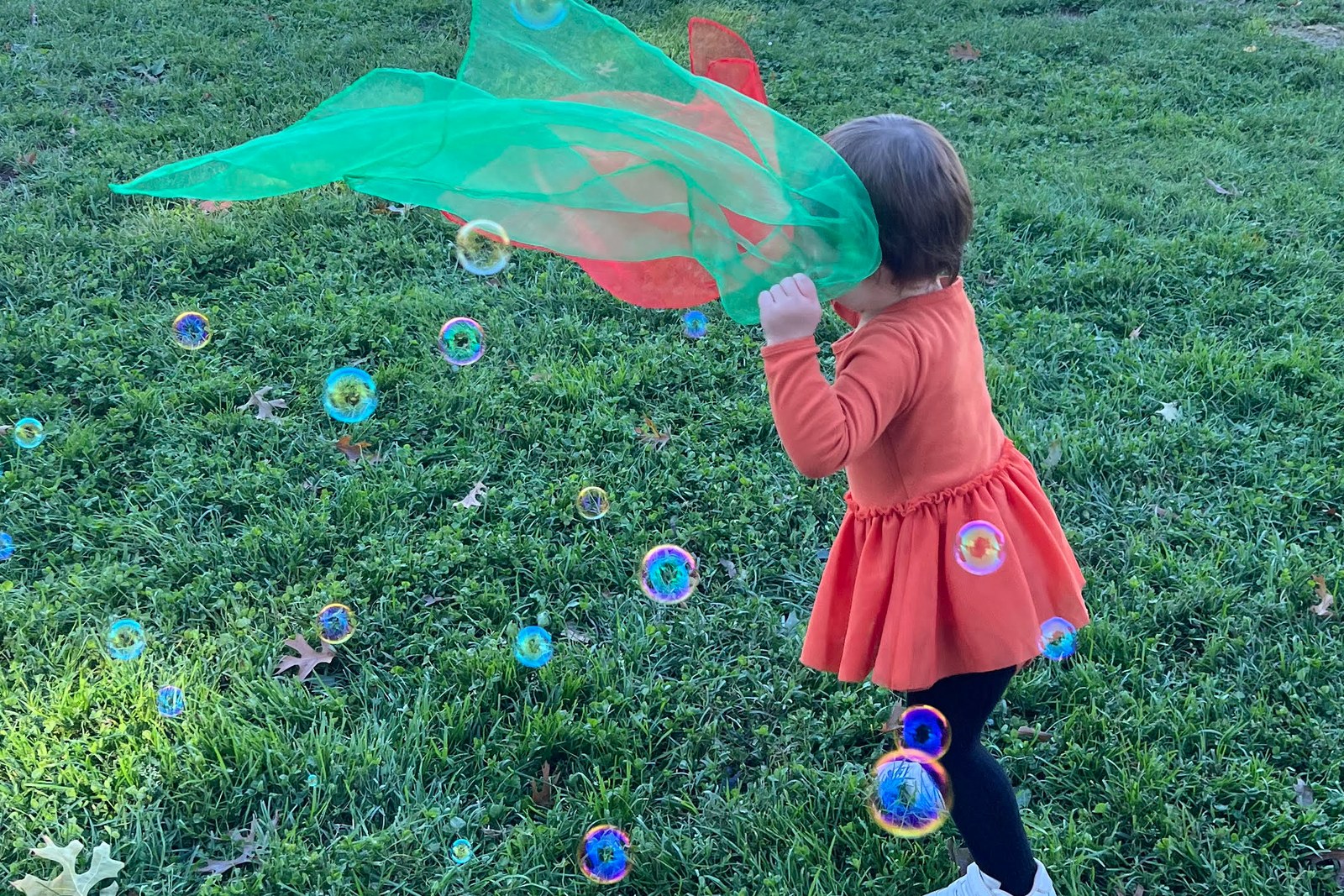 A three-year-old dancing in a park with a play silk and bubbles