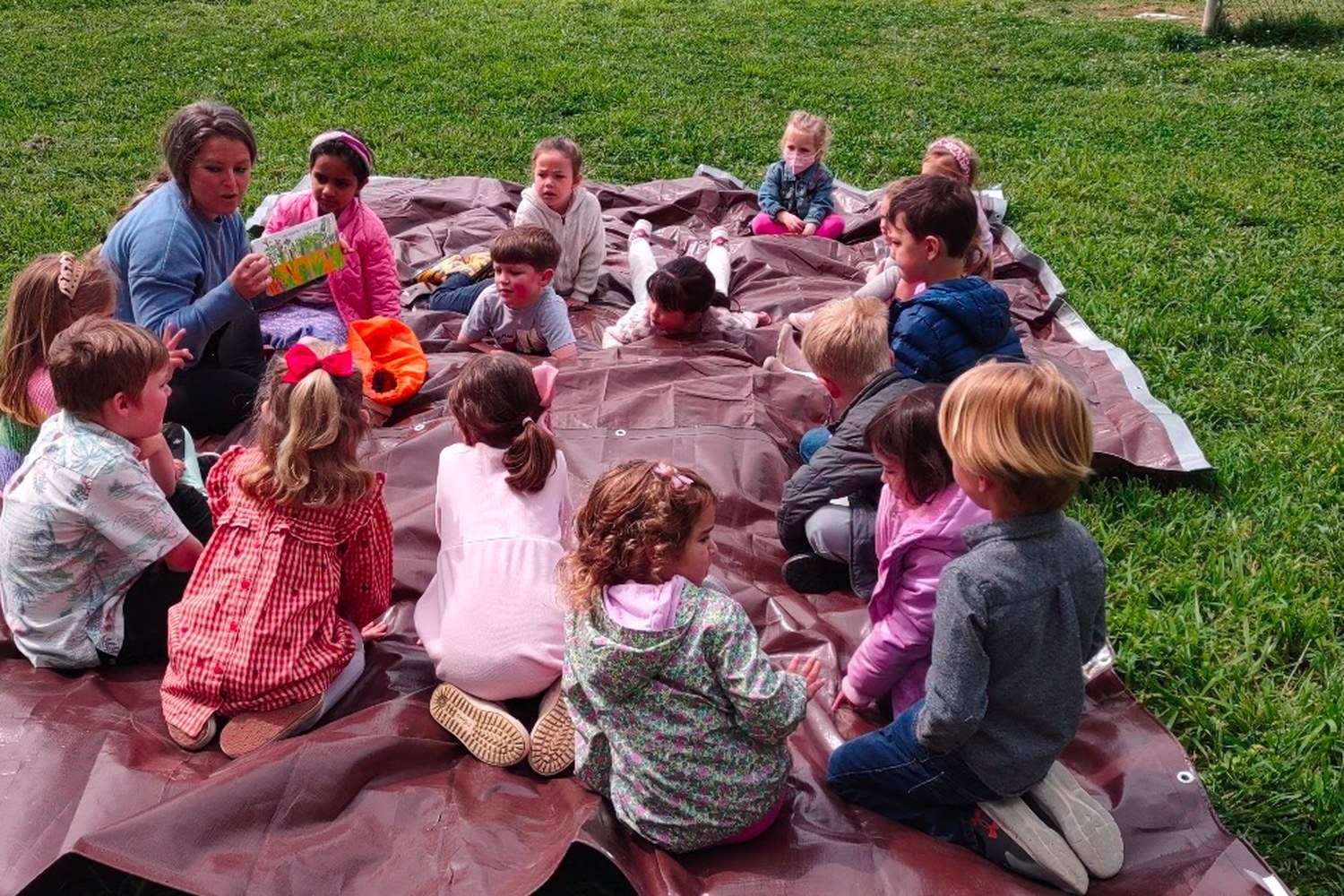 Chelsea reading and singing to a circle of preschoolers on a blanket outdoors