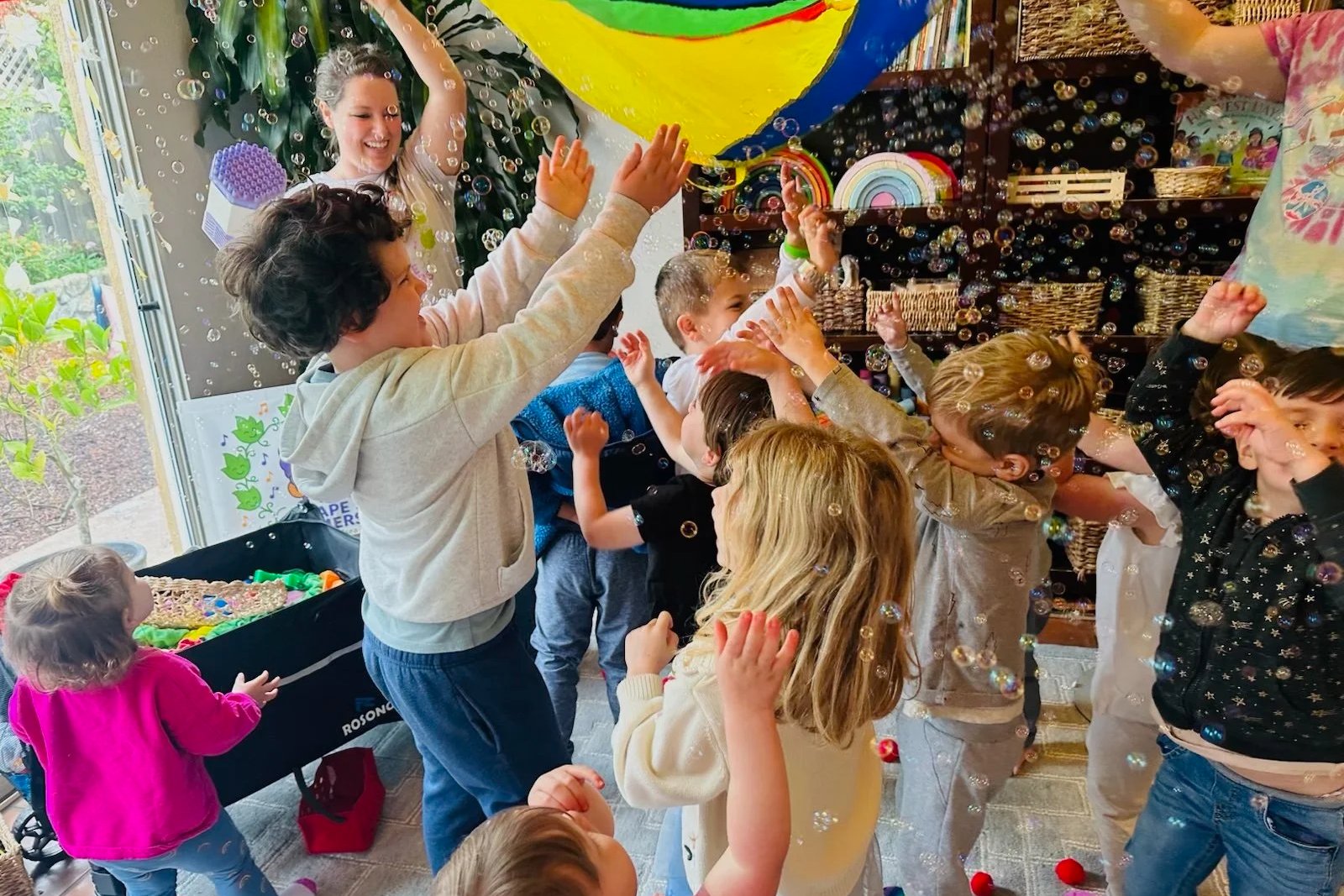 Chelsea leading a toddler music class, children reaching up to a rainbow parachute